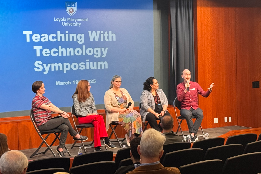 Photo of four panelist speakers (plus a moderator) sitting on a stage before an audience for a symposium event, with the words "Teaching with Technology" displayed on the screen behind them.
